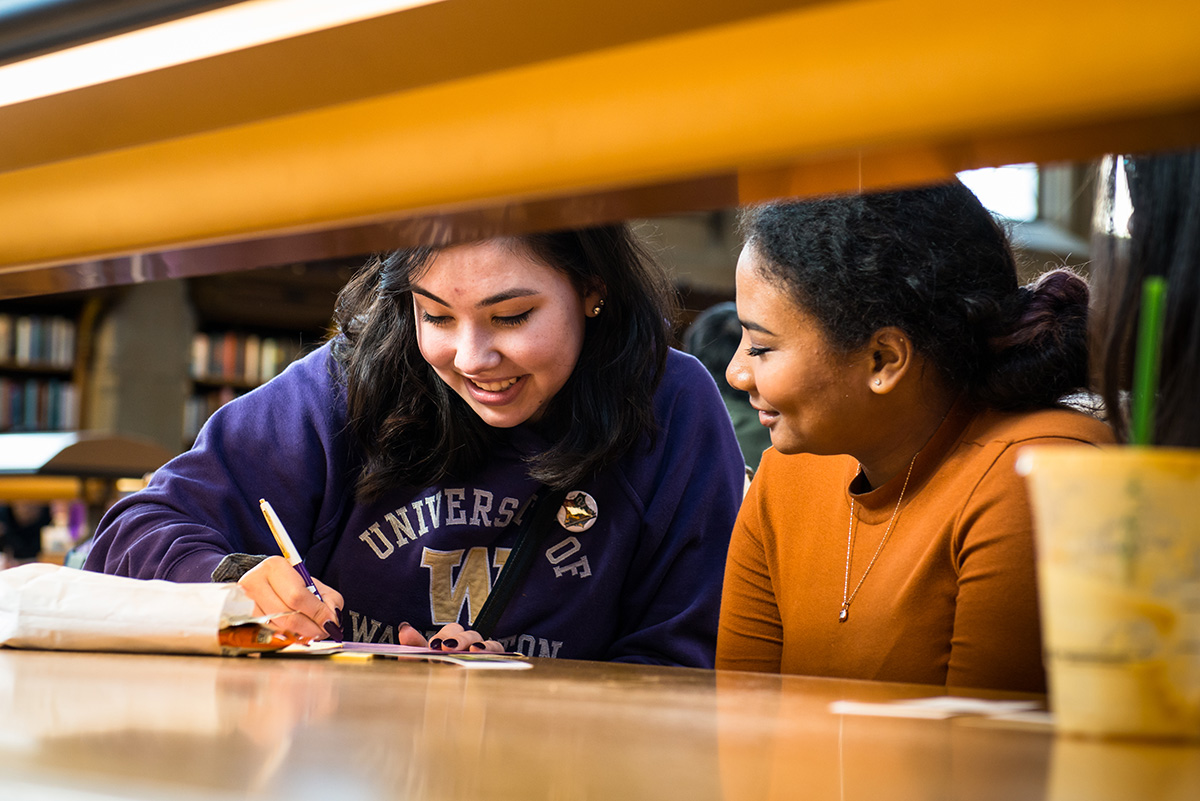 two students smiling and writing at a library table